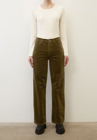Olivgrüne Cordhosen mit hoher Taille, geraden Beinen und Fronttaschen, kombiniert mit einem langen cremefarbenen Oberteil und schwarzen Schuhen.