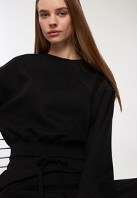 Maglione nero con vestibilità rilassata, colletto a costine e orlo elastico. Il tessuto sembra morbido, con maniche lunghe e dettagli minimi.