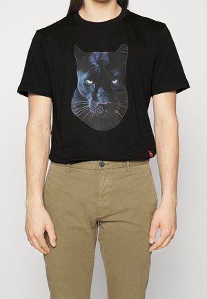 Hombre vistiendo una camiseta negra con un gran gráfico realista de la cara de una pantera negra en el pecho y pantalones beige.