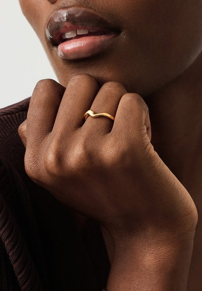 Κοντινό πλάνο χεριού με χρυσό κυματιστό δαχτυλίδι, ακουμπισμένο στο πηγούνι κοντά στα χείλη, που δείχνει σκούρο δέρμα και υφή καφέ υφάσματος στον καρπό.