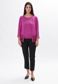 Fuchsiefarbener Cardigan mit Dreiviertelärmeln, filigranen Spitzenverzierungen am Saum, kombiniert mit einem passenden Oberteil und schwarzen, eng anliegenden Hosen sowie spitzen schwarzen Schuhen.