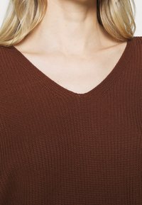 Maglione marrone lavorato a maglia con scollo a V, caratterizzato da un tessuto texturizzato in un'inquadratura ravvicinata, che mostra il motivo dei punti e il dettaglio del collo.