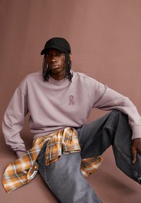 Sweatshirt violet avec un logo en ruban texturé, associé à un jean gris foncé. Une chemise à carreaux orange et gris est nouée autour de la taille. Casquette noire portée.