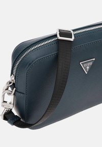 Bolso de hombro sintético de color azul marino con una superficie texturizada, cremallera plateada y placa triangular de marca; incluye una correa negra desmontable.