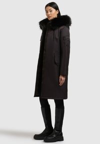 Sort parka med faux fur hætte, med lynlås og sidelommer, designet med en lang silhuet og en glat, blank tekstur.