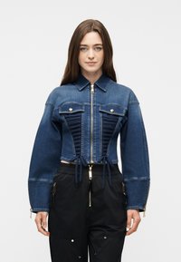 Γυναίκα που φοράει στενή μπλε τζιν μπουφάν με φερμουάρ μπροστά, με πιέτες στα πάνελ και μαύρο παντελόνι με χρυσές λεπτομέρειες φερμουάρ σε λευκό φόντο.