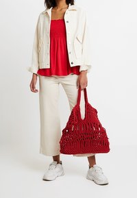 Röd vävd tote-väska med öppen design, ihop med en vit snörjacka över en röd smockad topp och krämfärgade vida byxor, avslutat med vita sneakers.