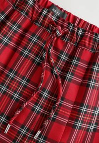 Pantalons de pyjama écossais à carreaux rouges, noirs et blancs avec taille élastique et cordon de serrage avec embouts en métal, étiquette portant la mention "NEXT" visible à l'intérieur.