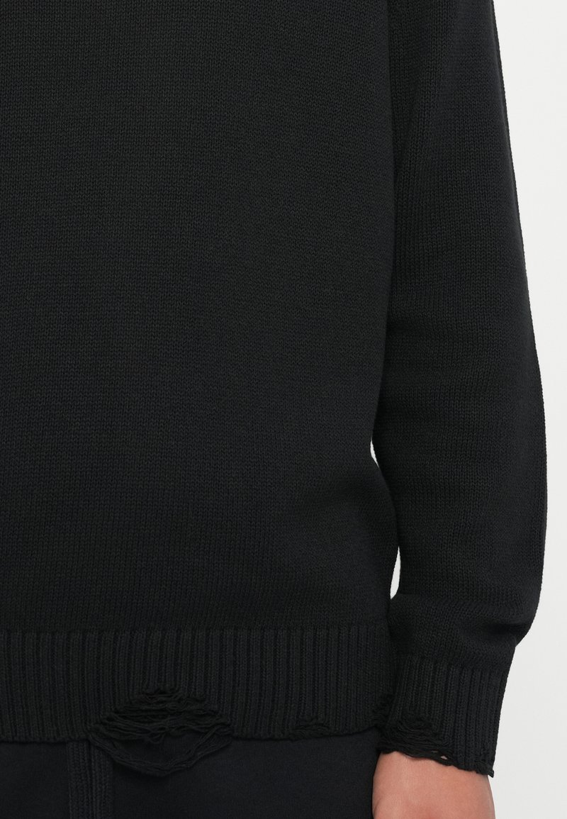 Zwarte gebreide trui met ribgebreide zoom en manchetten. Heeft een versleten gedeelte aan de onderkant, wat een textuurfinish benadrukt.