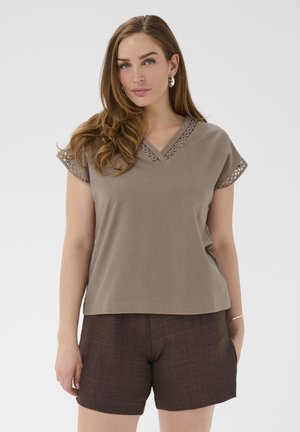 Pitkäaikahiuksinen nainen, jolla on taupen värinen V-kaula-aukko, jossa on silmäreikäreunus hihoissa ja kaula-aukossa, yhdistettynä tummanruskeisiin shortseihin.