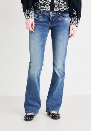 Frau trägt mittelblaue Bootcut-Jeans, schwarze Ballerinas mit Nieten und eine schwarze Jacke mit weißer Blumenstickerei.