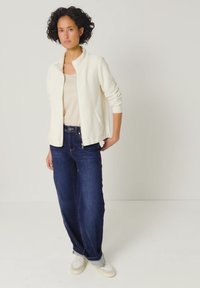 Frau mit lockigem Haar steht und trägt eine weiße Zip-Jacke, ein beiges Oberteil, dunkelblaue Jeans mit weitem Bein und weiße Sneakers vor einem einfarbigen Hintergrund.