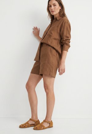 Kobieta ubrana w brązową koszulę na długi rękaw z guzikami, dopasowane spodenki i beżowe sandały z odkrytymi palcami, stojąca przy białej ścianie.