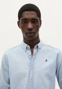 Lichtblauw overhemd met knoopsluiting en kraag, met een klein marineblauw doodskoplogo op de borst. Zachte stof met een gladde textuur.