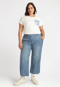 Camiseta blanca con un bolsillo de lentejuelas azules, combinada con unos pantalones vaqueros de denim azul claro de pierna ancha con detalles laterales. Zapatillas blancas completan el conjunto.