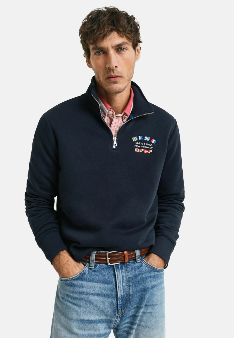 Pullover in pile blu navy con zip a metà, caratterizzato da loghi e bandiere ricamati. Indossato sopra una camicia a quadri rossi e bianchi, abbinato a jeans blu chiaro.