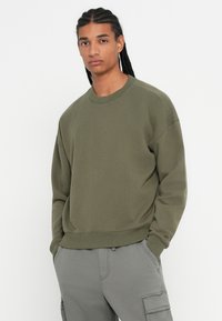 Giovane con capelli intrecciati che indossa una felpa verde oliva e pantaloni cargo grigio chiaro in piedi con le mani in tasca.