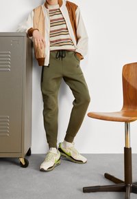 Pánský outfit zahrnující béžovo-hnědou bomber bundu, pruhované tričko, olivově zelené joggery s kapsami a světlé tenisky se zelenými akcenty.