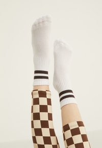 Calze alla caviglia bianche a coste con due strisce marroni in cima, abbinate a leggings check bianchi e beige. Lo sfondo è neutro.