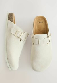 Des sabots en suède beige clair présentant une large sangle avec une boucle réglable, un double en mousse doux et une semelle en caoutchouc texturé pour l'adhérence.