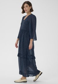 Marineblaue Maxikleid mit Polka-Dots, gestuften Rüschen, transparentem Stoff, V-Ausschnitt und Glockenärmeln, kombiniert mit hellen Loafers.