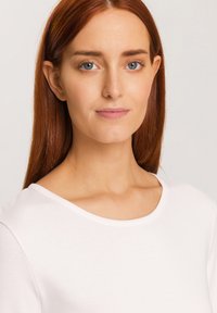 Bílý top s kulatým výstřihem, hladkou texturou a mírně přiléhavým střihem. Modelka s dlouhými kaštanovými vlasy jej nosí na neutrálním pozadí.