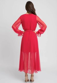 Czerwona sukienka w kropki z zwiewną, przezroczystą teksturą, długimi rękawami i ściągniętą talią. Spódnica sięga kostek i ma falbaniaste mankiety.