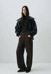 Veste en cuir noir avec un grand col, col roulé noir à pois, pantalon large marron et sandales à talons noires. Design minimaliste.