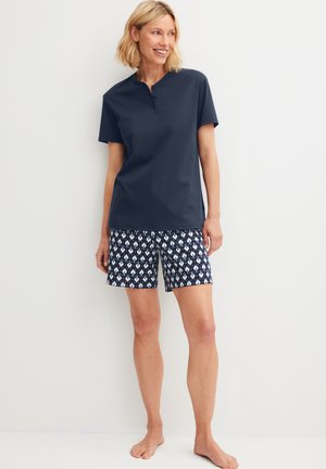 Woman smiling, standing barefoot against white wall, wearing navy short-sleeve henley shirt and navy patterned knee-length shorts.