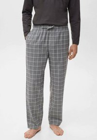 Uomo che indossa pantaloni da pigiama grigi a quadri con vita regolabile e una maglietta a maniche lunghe scura, in piedi a piedi nudi con le mani rilassate.