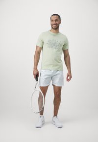 Hele roheline puuvillane T-särk, millel on graafiline disain, koos valgete triibuliste lühikesega ja valgete spordijalatsitega, hoides käes valget tennisereketit.