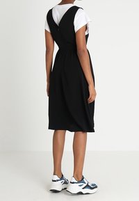 Černé bezramínkové šaty se střihem na míru v pase a volnou sukní, nošené přes bílé tričko s krátkým rukávem, doplněné bílými a modrými teniskami.