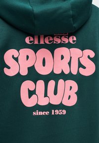 Σκούρα πράσινη μπλούζα με κουκούλα, με μεγάλο ροζ κείμενο "ellesse SPORTS CLUB since 1959" στην πλάτη κάτω από την κουκούλα.