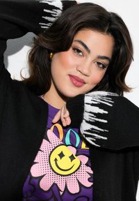 Cardigan noir avec un motif abstrait blanc sur les manches, porté sur un t-shirt violet affichant un graphique de fleur colorée avec un visage souriant.