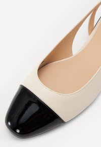 Ballerine en cuir beige avec un capuchon en cuir verni noir. Présente une texture lisse et une ouverture ronde pour l'entrée du pied.