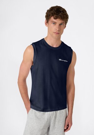 Giovane uomo con capelli ricci che indossa una maglietta senza maniche blu navy della Champion e pantaloncini grigio chiaro, in piedi con una mano in tasca.