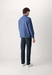 Lysblå jakke med krave, lange ærmer og lige snit, parret med mørke jeans og hvide sneakers, set fra ryggen.