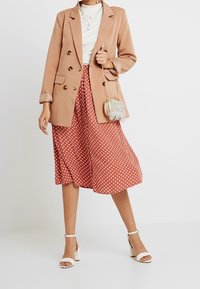 Blazer de doble botonadura marrón claro sobre una blusa blanca, combinado con una falda de longitud hasta la rodilla en color óxido con lunares blancos. Sandalias de tacón blancas.