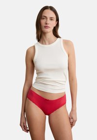 Frau trägt ein weißes ärmelloses geripptes Oberteil und rote Unterwäsche mit Spitzenbesatz, steht vor einem schlichten weißen Hintergrund.