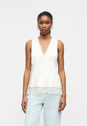 Femme aux cheveux tressés portant un haut blanc sans manches avec un col en V orné de dentelle et un jean bleu clair, debout devant un fond uni.