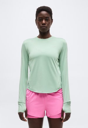 Kvinne står vendt fremover iført en mintgrønn langermet skjorte og skarpe rosa treningsshorts mot en ensfarget bakgrunn.