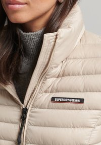 Béžová prošívaná bunda s vysokým límcem, vybavená zipperem a černou nášivkou s červenými akcenty. Šedý svetr viditelný pod ní.