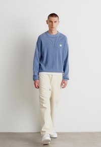 Sweater de malha oversized em azul claro, calças de cor creme, sapatilhas brancas; tecido texturado com um pequeno logo no peito.