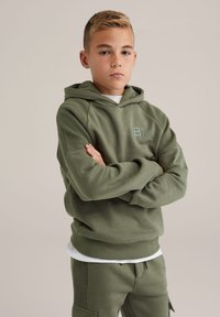 Olívazöld kapucnis pulóver, texturált mintával, elülső zsebbel és logóval a mellkasán, párosítva ugyanabból az anyagból készült nadrággal, oldalzsebekkel.