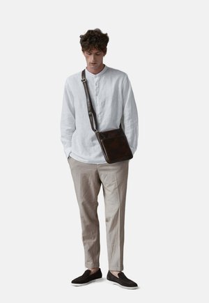 Witte linnen lange mouwen overhemd met een henley hals; beige maatpak broek; donkerbruine leren schoudertas; zwarte instap schoenen.