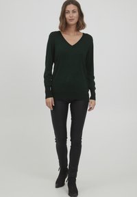 Mørkegrøn, let sweater med V-hals og lange ærmer, båret med sorte, tætsiddende bukser og ankelstøvler. Enkel, glat tekstur.