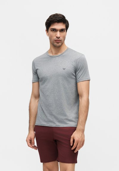 Άνδρας με γκρι κοντομάνικη μπλούζα με λογότυπο στο στήθος και μπορντό σορτς, στέκεται μπροστά σε λευκό φόντο.