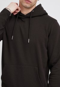 Hoodie marron foncé en tissu doux, avec une poche avant, une capuche à cordon de serrage avec œillets en métal, et une coupe décontractée.