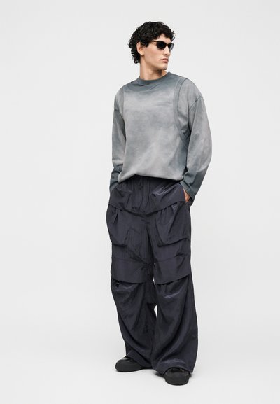 Άνδρας που φορά γκρι μπλούζα με μακριά μανίκια, φαρδιά μαύρα παντελόνια cargo με μεγάλες τσέπες, μαύρα παπούτσια και γυαλιά ηλίου, στέκεται με τα χέρια στις τσέπες.