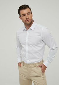 Chemise blanche à col écarté, coupe slim et manches longues, accompagnée de pantalons beiges. Texture lisse, sans motifs visibles.